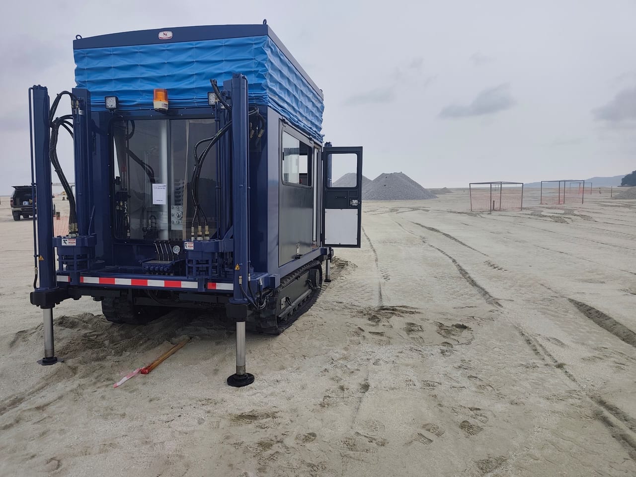 Blue enclosed crawler CPT rig on sandy ground, viewed from the hydraulic end with the side door open and support legs lowered