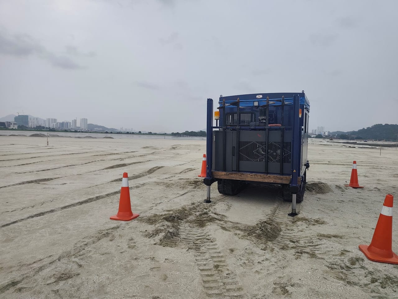Ventilated end of a blue crawler CPT rig on sandy ground, with orange traffic cones and track marks in the foreground