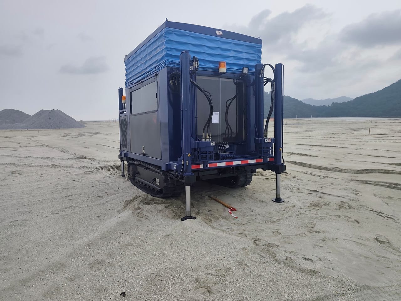 Three-quarter view of a blue crawler CPT rig showing its closed side panel, hydraulic hoses, tracks and lowered support legs